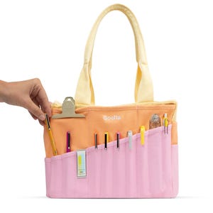 May include: A peach-coloured tote bag with a pink front pocket, holding various pens and pencils. The bag has a yellow handle and a metal clip. The word "Soella" is printed on the front.