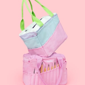 Könnte beinhalten: Zwei rosa Stofftaschen mit Henkeln. Die obere Tasche ist hellblau, lavendelfarben und weiß mit einem grünen Henkel. Die untere Tasche enthält verschiedene Werkzeuge. Beide Taschen haben das Wort "Scolia" aufgestickt.