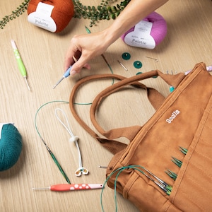 Pode incluir: Uma bolsa tote de lona marrom com a palavra "Spoola" bordada na frente. A bolsa tem vários bolsos e está cheia de agulhas de tricô. Também há vários materiais de tricô espalhados pela bolsa, incluindo lã, marcadores de ponto e agulhas de crochê.