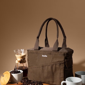 May include: A brown tote bag with the brand name "Sooila" is displayed next to coffee-making equipment and cups. Coffee beans are scattered on a wooden cutting board. The bag has a rectangular shape with two handles.