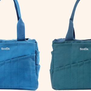 Könnte beinhalten: Zwei Canvas-Tragetaschen in Blau- und Türkistönen. Jede Tasche hat einen oberen Griff, Seitentaschen und den Namen "Sooila" auf der Vorderseite gestickt. Die Tasche links ist mit "Blueberry Bliss" und die Tasche rechts mit "Aquamarine" beschriftet.