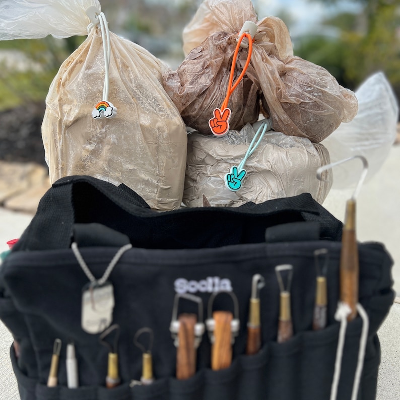 May include: A black tool bag with the word "Sculptia" embroidered on the front. The bag is filled with various sculpting tools and has a silver chain with a tag hanging from the top. The bag is sitting on a table with two plastic bags in the background. The plastic bags have peace sign and rainbow stickers on them.
