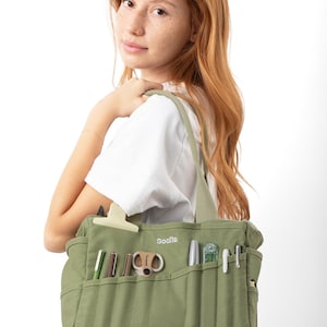 A young woman with red hair is holding a green tote bag with various items, including pens and scissors, in it.