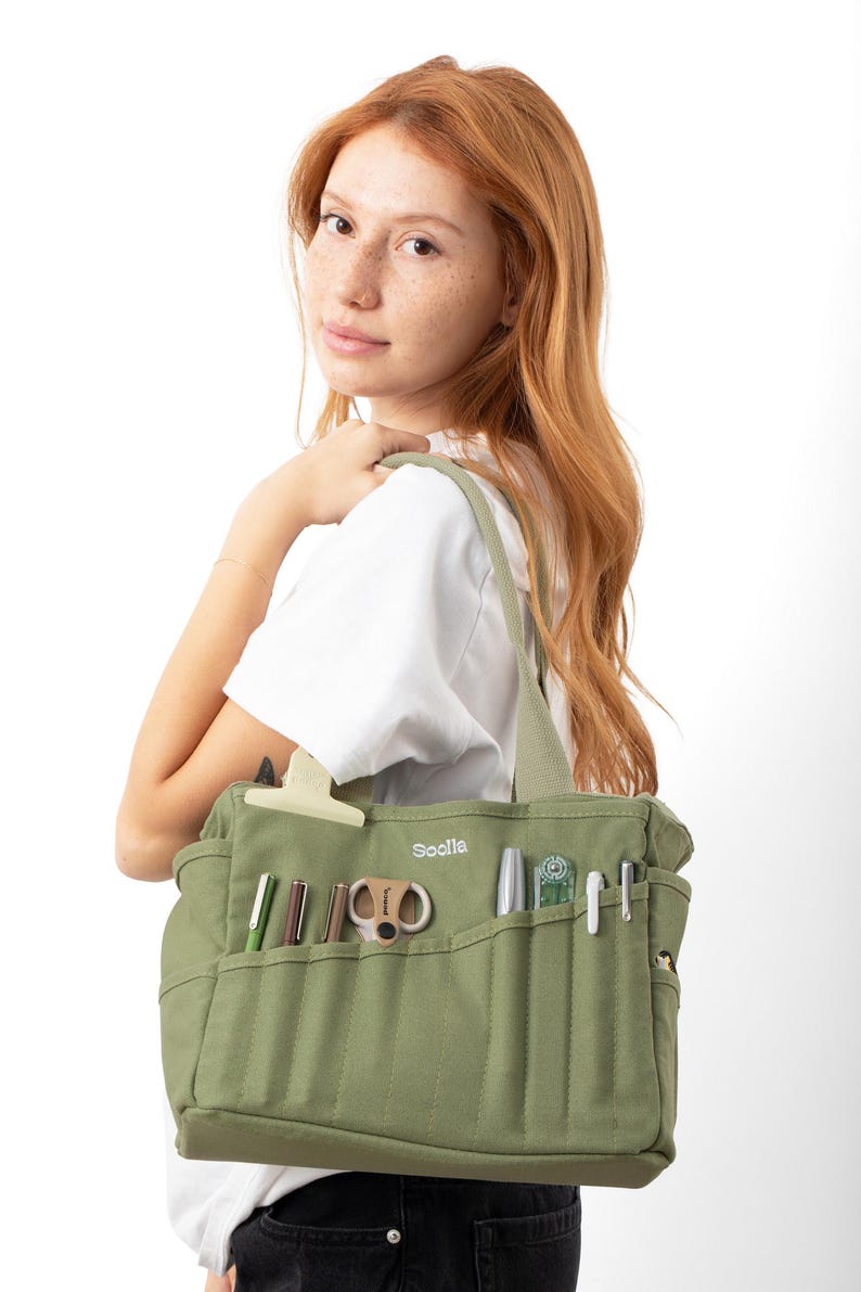 A young woman with red hair is holding a green tote bag with various items, including pens and scissors, in it.