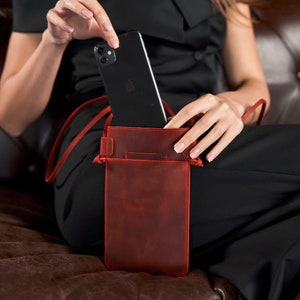 May include: A red leather phone case with a strap. The case has a flap that closes with a snap. A black smartphone is inside the case.