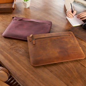 May include: Two leather laptop sleeves, one brown and one burgundy, with zipper closures. The sleeves are lying on a wooden surface.