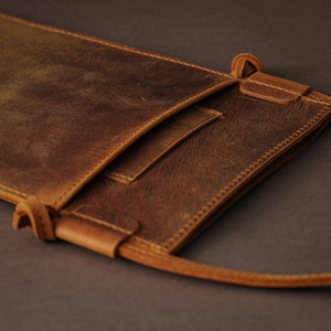 May include: A brown leather phone pouch with a flap closure and a long strap. The pouch has a distressed look and is made of high-quality leather.