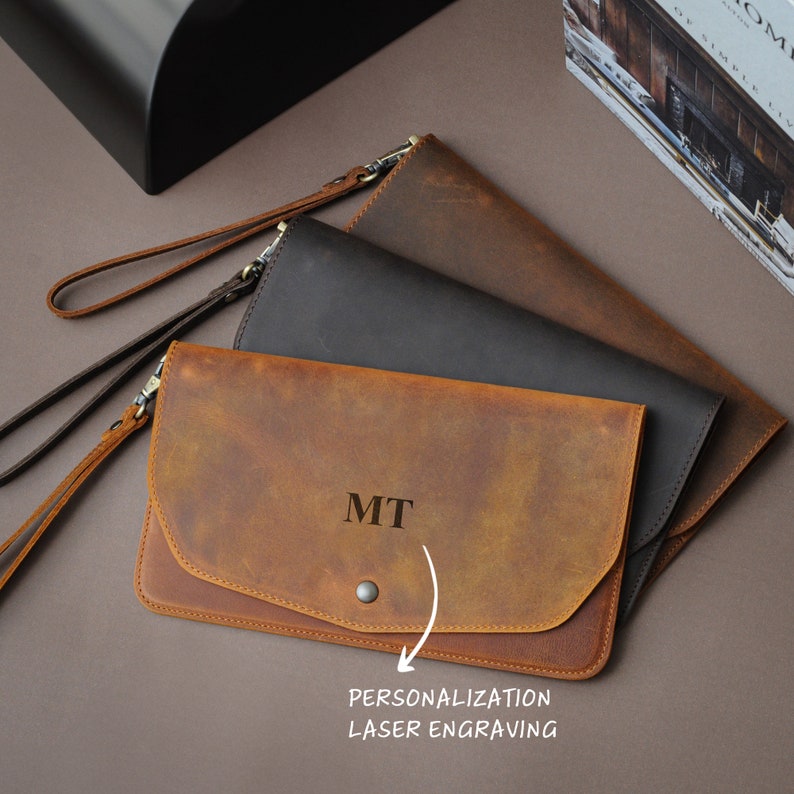 May include: Three leather wallets in brown, black, and tan. The tan wallet has the initials "MIT" engraved on the front. The wallets have a flap closure and a wrist strap.