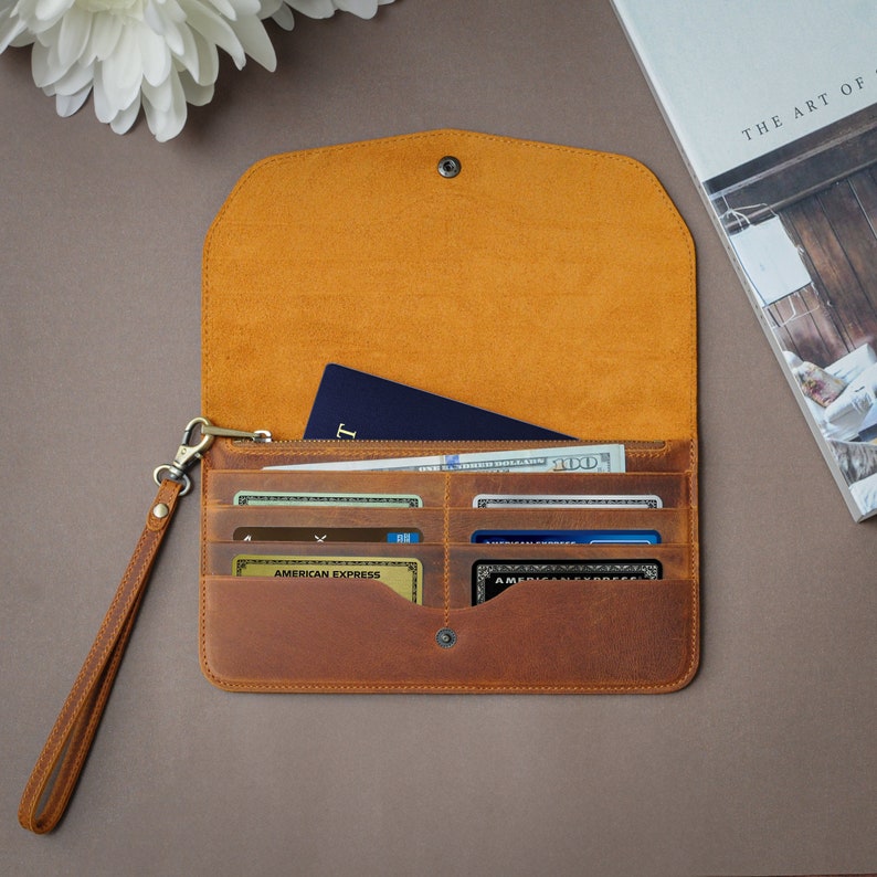 May include: A brown leather wallet with a flap closure, a wrist strap, and multiple card slots. The wallet is open and contains a passport, credit cards, and cash.