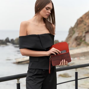 May include: A woman wearing a black off-the-shoulder top and black pants holds a red leather wallet with a black phone inside.