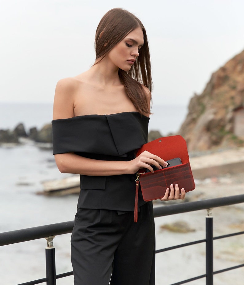 May include: A woman wearing a black off-the-shoulder top and black pants holds a red leather wallet with a black phone inside.