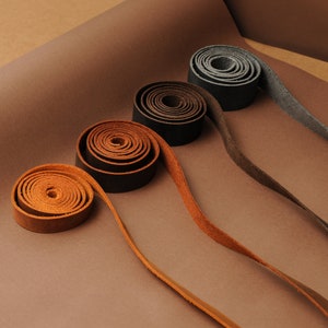 May include: Four leather straps of different colors and widths are rolled up and laid out on a brown surface. The straps are brown, dark brown, gray, and black.