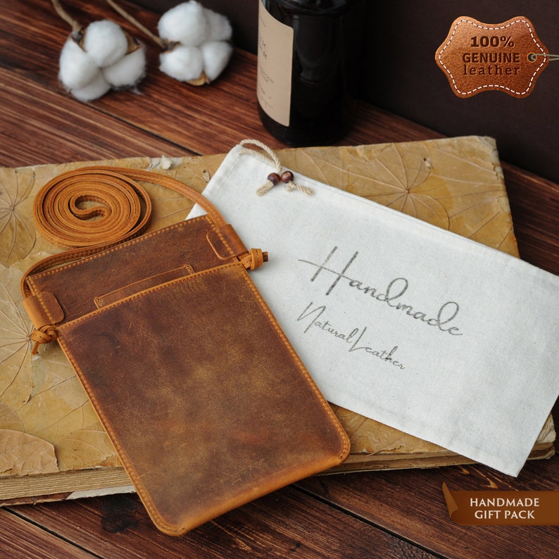 May include: A brown leather phone pouch with a flap closure and a long strap. The pouch is sitting on a wooden surface with a white cloth bag that says "Handmade Natural Leather" and a brown leather tag that says "100% Genuine Leather".