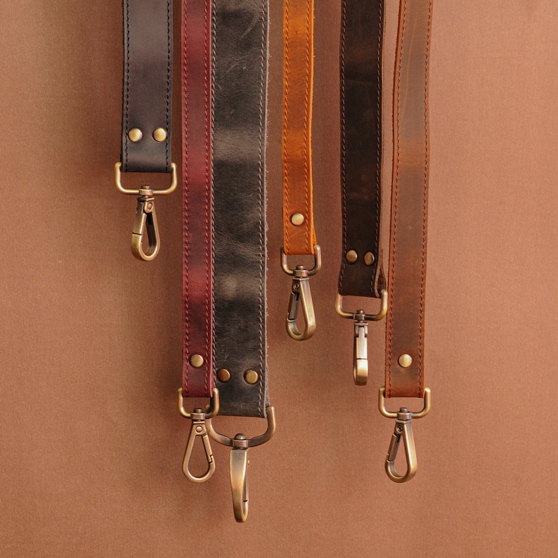 Op de afbeelding: Een verzameling leren riemen in diverse kleuren, waaronder zwart, bordeauxrood, grijs, oranje en bruin. Elke riem is voorzien van messingkleurige metalen hardware, waaronder klinknagels en draaibare haken. De riemen zijn ontworpen voor tassen of andere accessoires.
