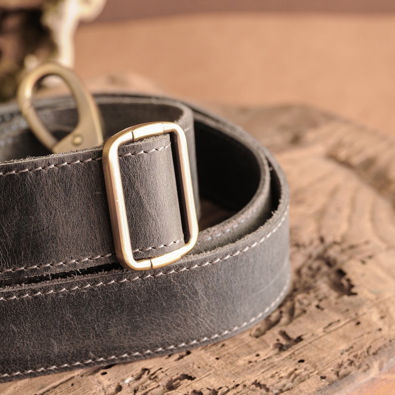 Op de afbeelding: Close-up van een donkergrijze leren riem met witte stiksels en messingkleurige hardware. De riem is opgerold en heeft een rechthoekige gesp en een draaibare sluiting. Het leer heeft een getextureerde uitstraling.
