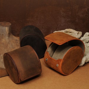 May include: Three leather watch boxes in brown and dark brown. The boxes are cylindrical and have a hinged lid. The boxes are made of a soft, supple leather.