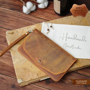 May include: A brown leather wallet with a flap closure and a wrist strap. The wallet is made of genuine leather and is handmade. The wallet is on a wooden surface with a white cloth bag and a cotton ball in the background. The text "Handmade Natural Leather" is printed on the cloth bag. The text "100% Genuine Leather" is printed on a brown leather tag.