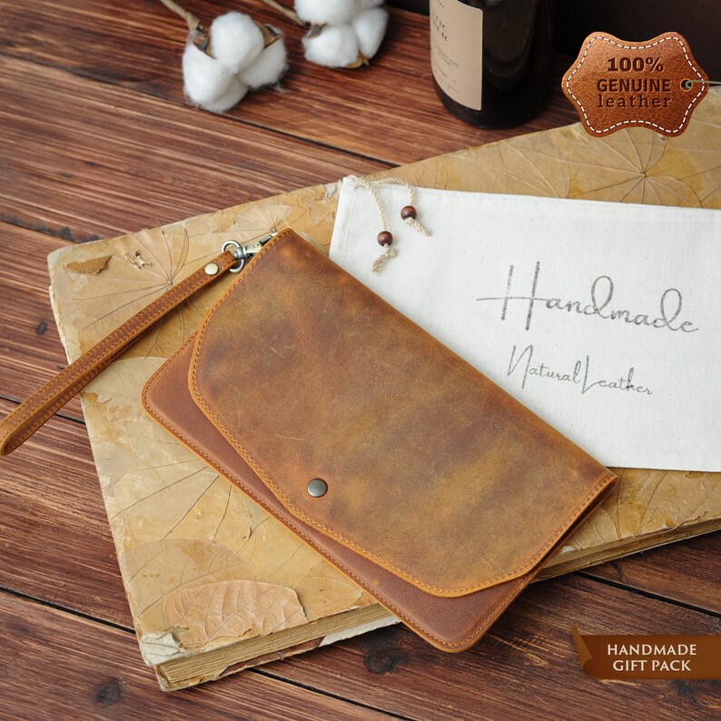 May include: A brown leather wallet with a flap closure and a wrist strap. The wallet is made of genuine leather and is handmade. The wallet is on a wooden surface with a white cloth bag and a cotton ball in the background. The text "Handmade Natural Leather" is printed on the cloth bag. The text "100% Genuine Leather" is printed on a brown leather tag.