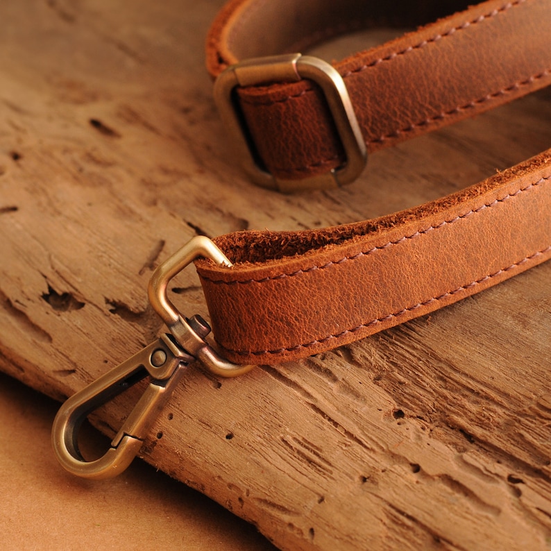 Op de afbeelding: Close-up van een bruine leren riem met een messingkleurige metalen sluiting en gesp. De riem is gestikt met een lichtere bruine draad. De achtergrond is een houten oppervlak met textuur.