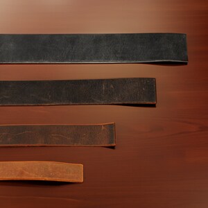 Puede incluir: Cuatro correas de cuero de diferentes anchos y colores, incluyendo negro, marr&oacute;n y tostado, se colocan sobre una superficie de madera.