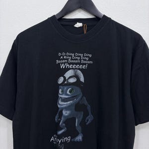 May include: A black short-sleeved t-shirt featuring a graphic of a blue creature wearing goggles. The text reads: D-D-Ding Ding Ding A Ring Ding Ding Baaam Baaam Baaam Wheeeee! and the Anoying Thing at the bottom. The shirt is on a black hanger.