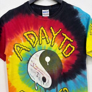 Puede incluir: Una camiseta de manga corta con estampado tie-dye en espiral en rojo, amarillo, azul y negro. El frente muestra un símbolo yin-yang que se derrite con el texto amarillo A DAY TO REMEMBER arqueado sobre y debajo del gráfico en cuello negro.