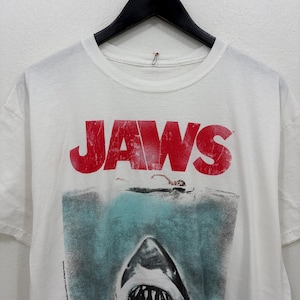 May include: A white short-sleeved t-shirt featuring the red Jaws movie logo text above a graphic of a shark swimming towards a person floating on the water surface. The design includes a small copyright notice printed vertically on the lower left side.