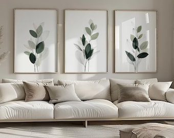 May include: Three framed botanical prints with green and gray leaf designs hang above a cream-colored sofa with several pillows. The artwork features a minimalist style, adding a touch of nature to the room's decor.