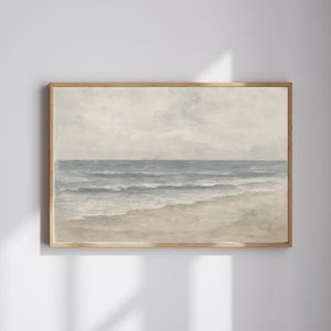 May include: A framed landscape painting of a beach scene. The artwork depicts a serene ocean with gentle waves, a sandy shore, and a cloudy sky. The color palette is soft, featuring muted blues, beige, and gray tones, creating a calming aesthetic.