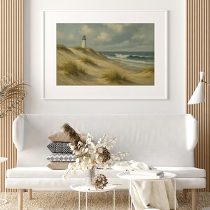 May include: Framed landscape art featuring a lighthouse on a sandy dune, with ocean waves under a cloudy sky. The artwork is displayed above a white sofa with decorative pillows and small white side tables. The room has a neutral color palette.