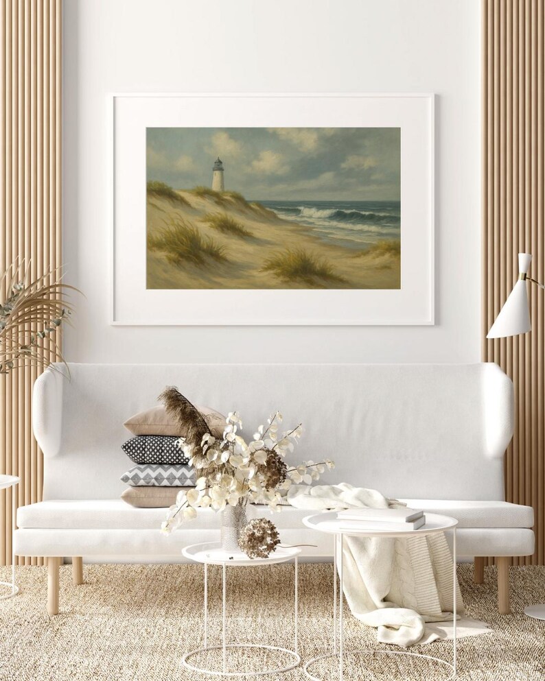 May include: Framed landscape art featuring a lighthouse on a sandy dune, with ocean waves under a cloudy sky. The artwork is displayed above a white sofa with decorative pillows and small white side tables. The room has a neutral color palette.