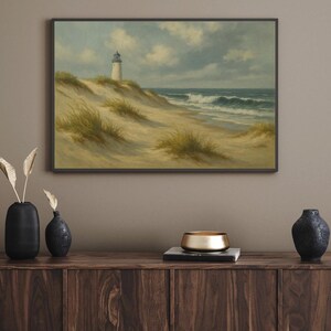 May include: A framed landscape painting depicts a lighthouse on a sandy dune overlooking the ocean. The artwork features a white lighthouse, tan sand, green grass, and a blue sky with clouds. The painting is displayed above a wooden cabinet.