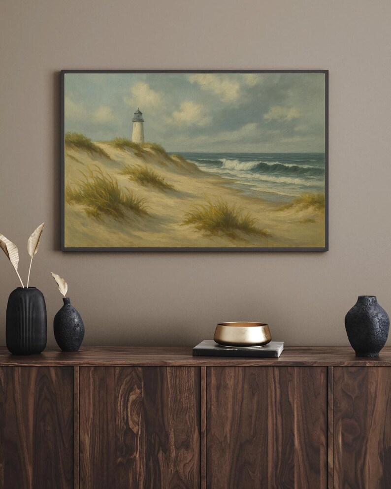 May include: A framed landscape painting depicts a lighthouse on a sandy dune overlooking the ocean. The artwork features a white lighthouse, tan sand, green grass, and a blue sky with clouds. The painting is displayed above a wooden cabinet.