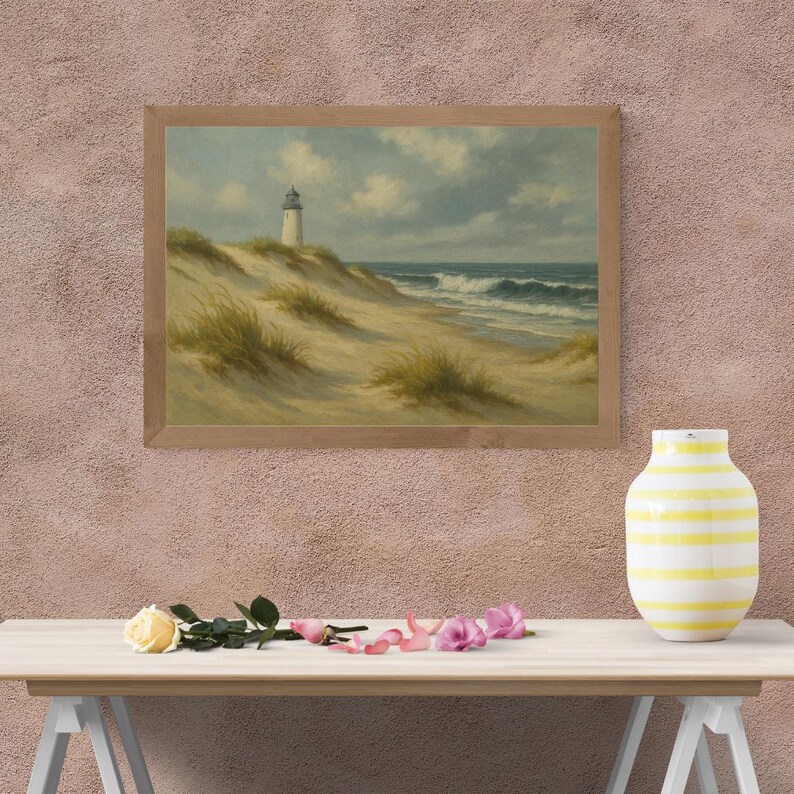 May include: A framed landscape painting depicts a lighthouse on a sandy dune overlooking the ocean. The artwork features a light blue sky with clouds, waves, and beach grass. The frame is made of light-colored wood. A yellow and white striped vase sits on a table.