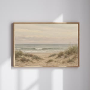 May include: A framed landscape painting depicts a serene beach scene. The artwork features a sandy path leading to the ocean, with dunes and grasses in the foreground. The sky is overcast, and the overall color palette is muted with beige, green, and blue tones.