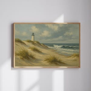 May include: A framed landscape painting depicts a coastal scene. A white lighthouse stands atop a sandy dune, with ocean waves crashing on the shore. The sky is overcast with clouds, and the artwork is framed in a light wood.