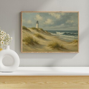 May include: A framed landscape painting depicts a coastal scene with a white lighthouse atop a sand dune. The artwork features a sandy beach, ocean waves, and a cloudy sky. The painting is in a wooden frame and evokes a serene, nautical theme.