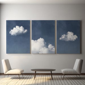 May include: A triptych of framed cloudscape prints in a living room setting. Each canvas displays a white cloud against a dark blue backdrop. The room includes two white chairs and a dark oval coffee table.