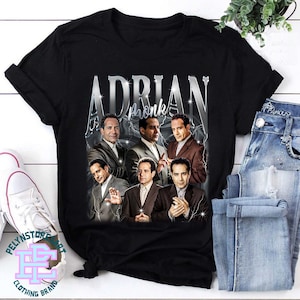 Peut inclure: T-shirt noir avec un graphique argenté d'Adrian Monk et le texte "Adrian Monk" dans une police argentée.