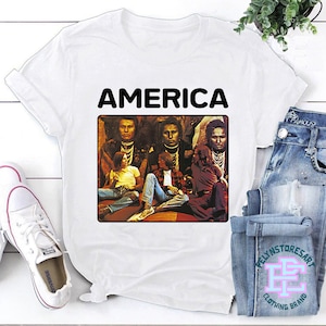 Può includere: Maglietta bianca con una stampa nera della copertina dell'album "America" di America. La copertina dell'album presenta un gruppo di quattro persone sedute su un tappeto con uno sfondo marrone.