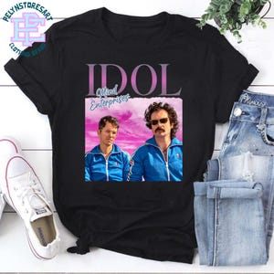 Può includere: T-shirt nero con la scritta "IDOL Mind Enterprises" in rosa e bianco. La maglietta mostra l'immagine di due uomini in tuta blu. Sono visibili anche scarpe da ginnastica bianche e jeans blu.