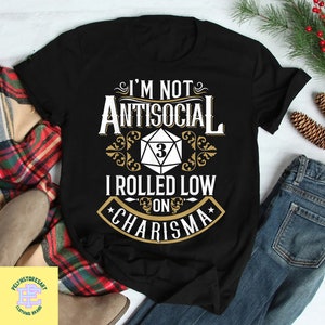 Könnte beinhalten: Schwarzes T-Shirt mit einer weißen Grafik, auf der "Ich bin nicht unsozial, ich habe niedrig auf Charisma gewürfelt" steht, mit einem goldenen Würfel mit einer 3 darauf.