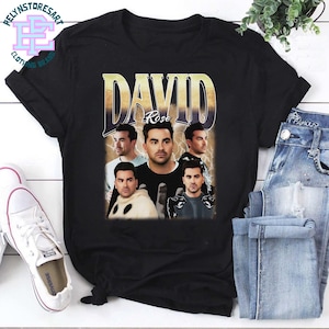 May include: Black t-shirt featuring a vintage-style graphic of a man with the name "DAVID" in large letters and "Rose" underneath. The graphic includes multiple images of the man. The shirt is paired with blue jeans and white sneakers.