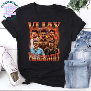 Op de afbeelding: Zwart T-shirt met een afbeelding van een man en de tekst "VIJAY THALAPATHY" in oranje. Het shirt wordt gecombineerd met een blauwe spijkerbroek en witte sneakers.