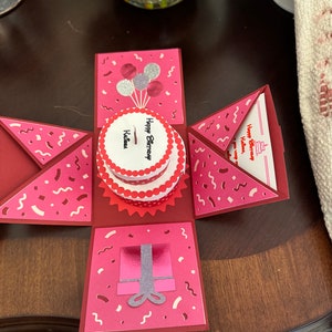 May include: A handmade, open birthday card with a red and pink color scheme. The card features a pop-up cake, balloons, and a gift box. The words "Happy Birthday" and a name are visible on the cake.