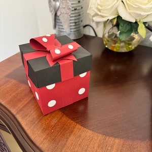 以下が含まれることがあります： 黒い上部と赤いベースの装飾的なギフトボックスで、白い水玉模様で飾られています。ボックスは、白い水玉模様の赤いリボンで結ばれています。ボックスは、暗い木製の表面に置かれています。