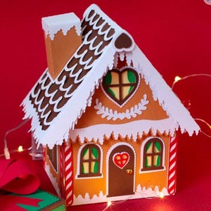 以下が含まれることがあります： 茶色の屋根、白いトリム、複雑なディテールが施された、装飾的なジンジャーブレッドハウス。 ハート型の窓とドアがあり、赤と白のストライプのアクセントが付いています。 赤い背景に、ギフトボックスとフェアリーライトが配置されています。