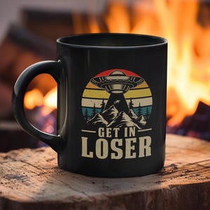 May include: Black ceramic coffee mug with a retro design featuring a flying saucer, mountains, and the text "Get in Loser".