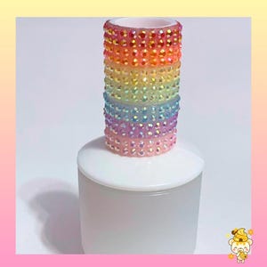 May include: A rainbow-colored, bedazzled cylinder sits atop a white, frosted glass bottle. The cylinder is covered in small, iridescent rhinestones, transitioning through shades of red, orange, yellow, green, blue, and purple. The bottle has a rounded top.