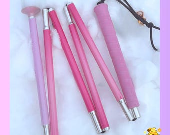 May include: A collapsible mobility cane featuring a light pink shaft, a darker pink textured grip, and a rounded white tip. The cane is segmented into multiple sections connected by internal cords, with a black wrist strap attached to the handle.